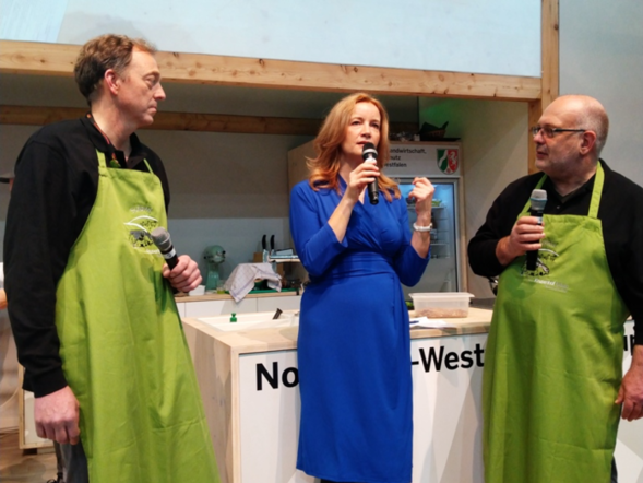 2 Männer mit grüner Kochschürze, auf der Bühne von der grünen Woche. In der Mitte die Moderatorin im blauem Kleid und Mikro