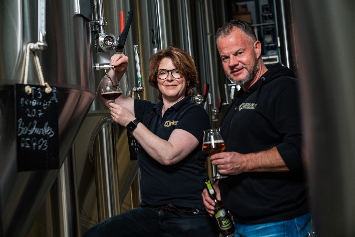 Eine Frau und ein Mann mit schwarzen T-Shirts stehen vor einem Braukessle für Bier. Die Frau zapft aus dem Braukessel Bier in ein Glas